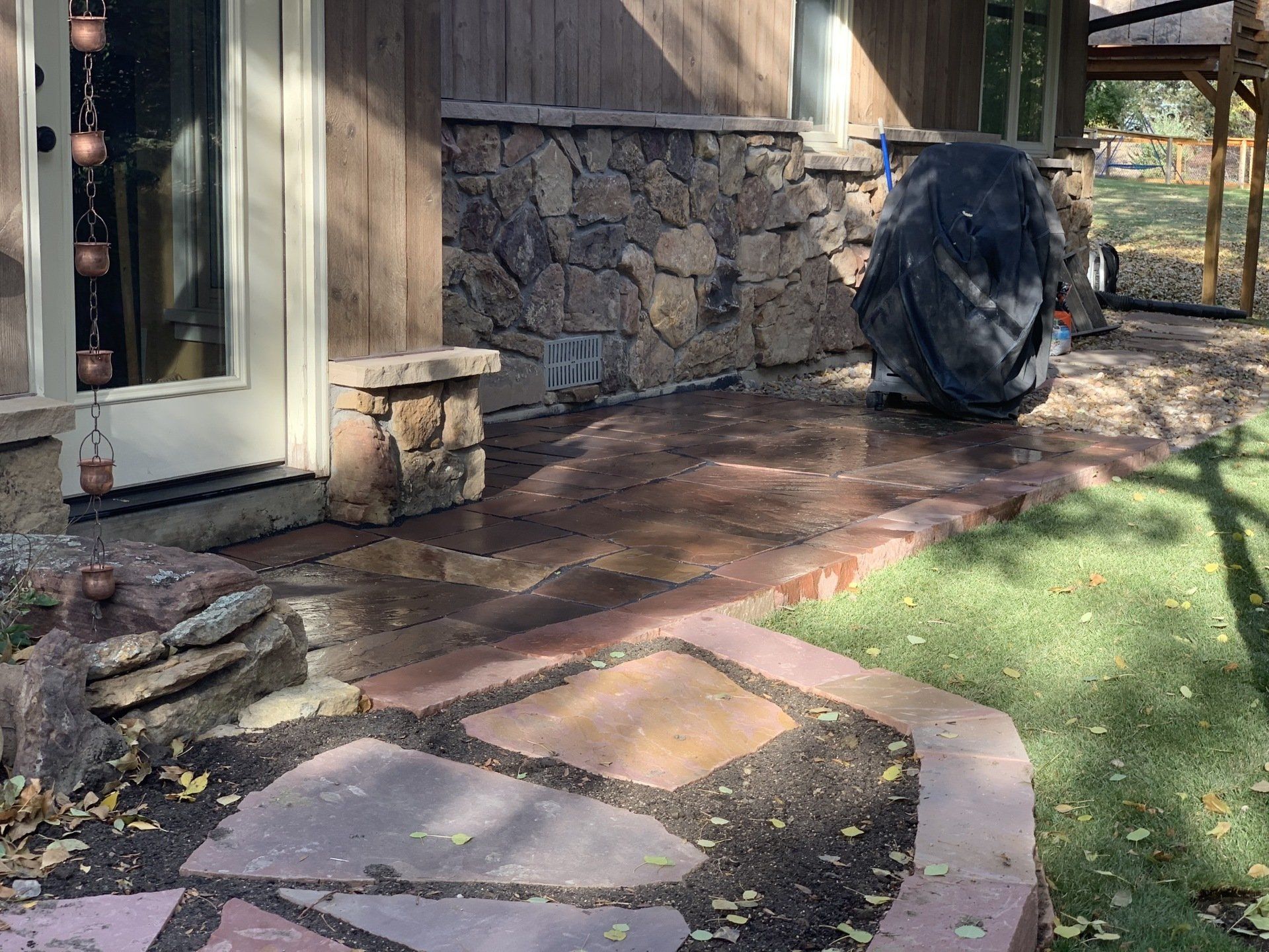 Stone patio and walkway leading to a house with a grill covered by a black cover and grass.