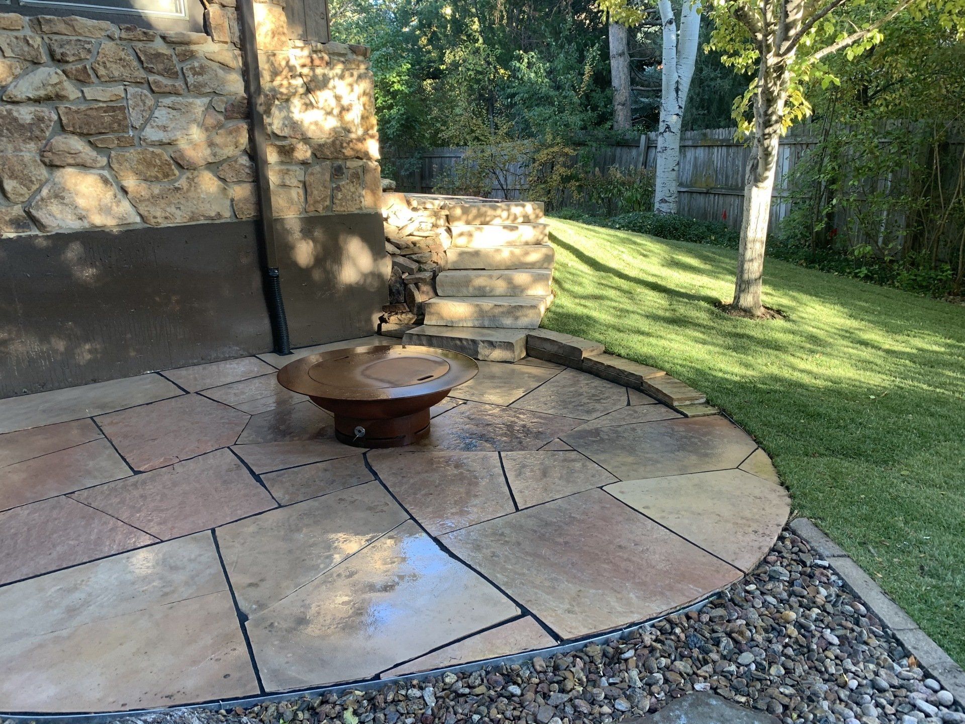 A backyard patio with a fire pit, bordered by grass and a stone wall.