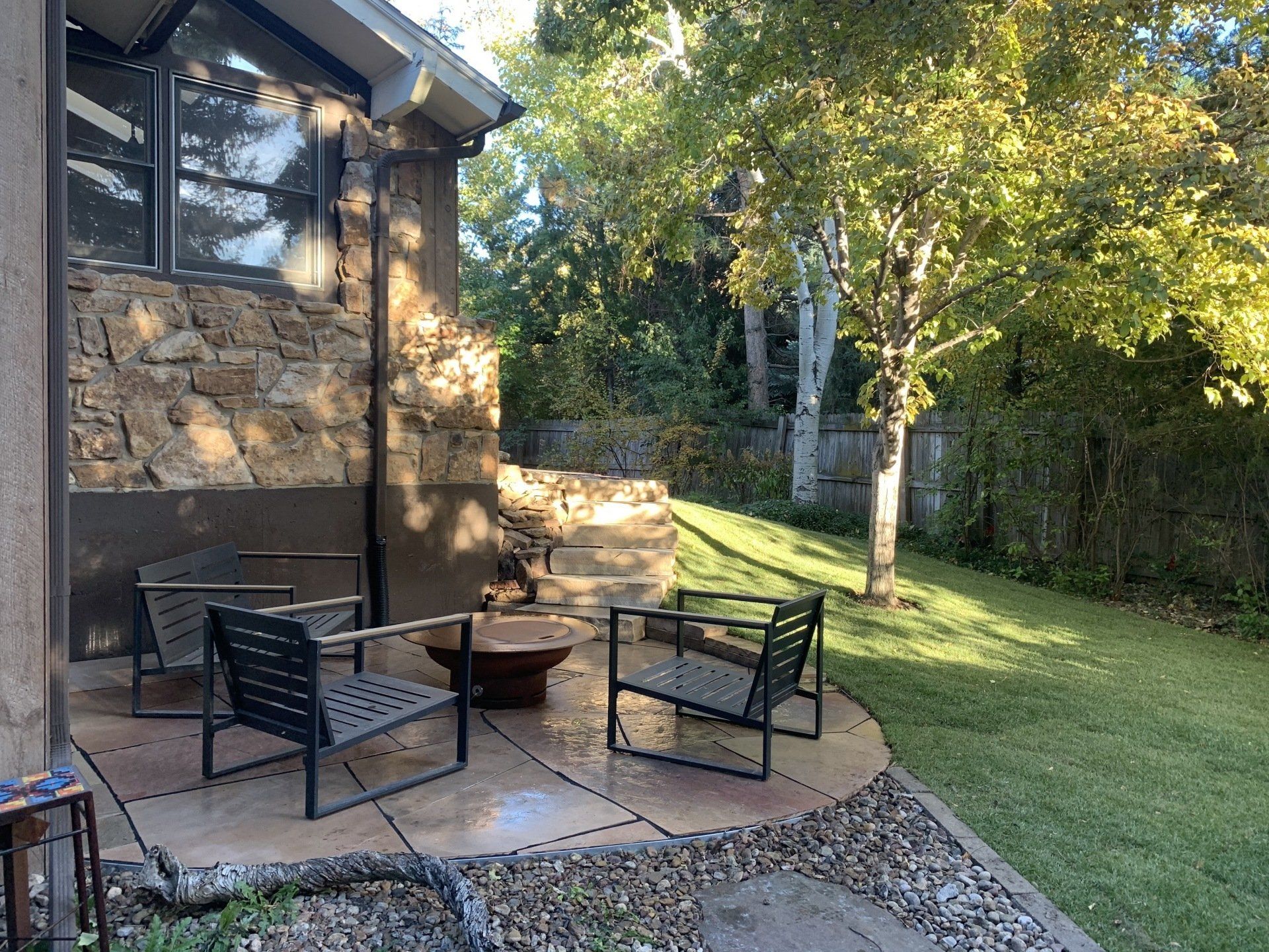 Patio with chairs, fire pit, and stone wall, surrounded by grass and trees.