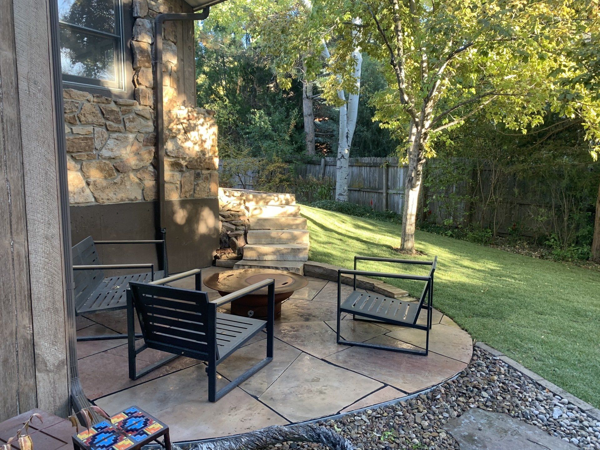 Patio with two modern chairs, fire pit, stone wall, and grassy yard.