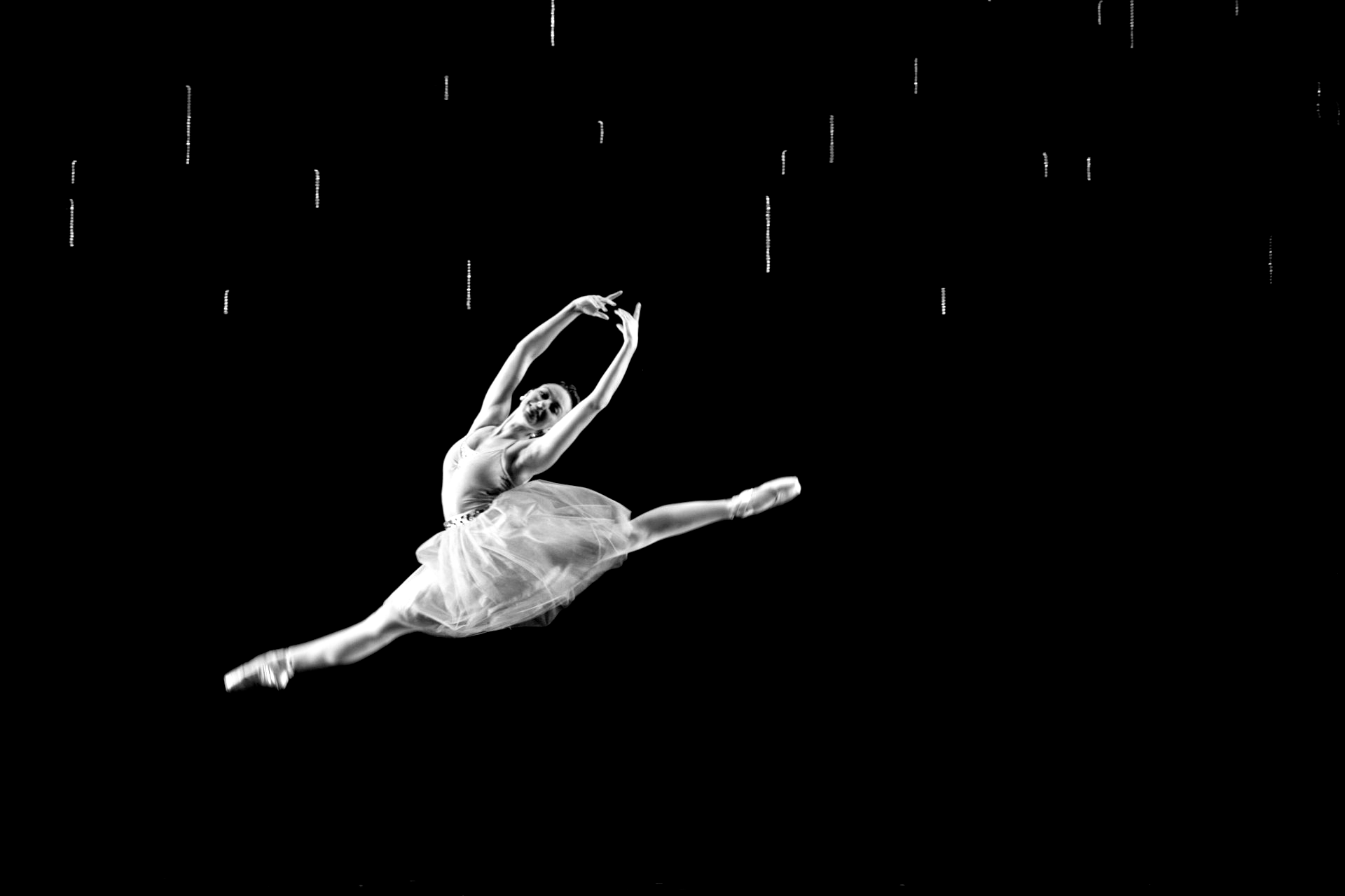 Ballerina leaps mid-air, legs split, arms overhead. Black and white with suggested falling rain.