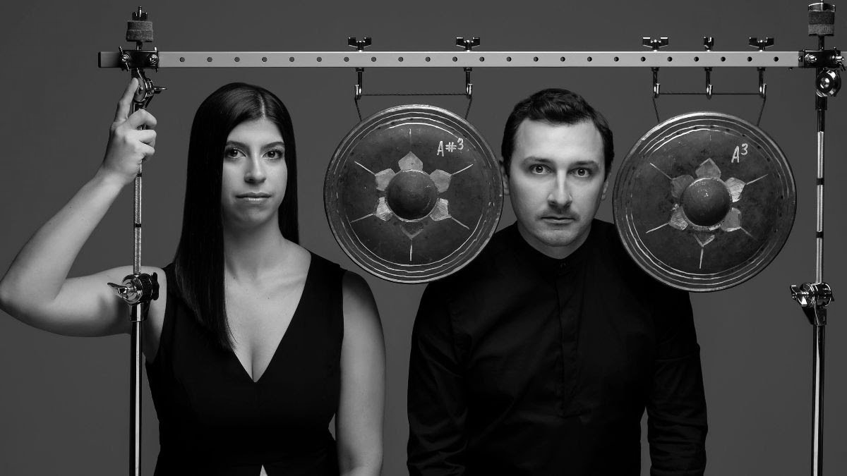 Woman and man in black attire, flanking large gongs on a metal frame. Woman points up. Black and white.