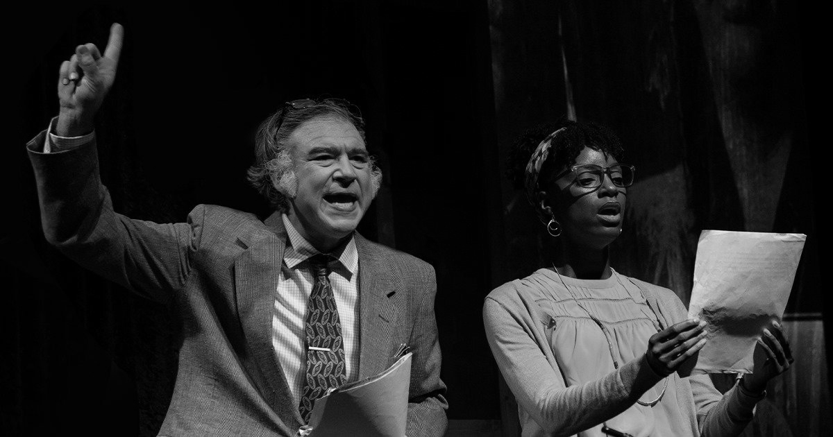 A man and a woman on stage, reading from papers. The man gestures upwards; both are singing.