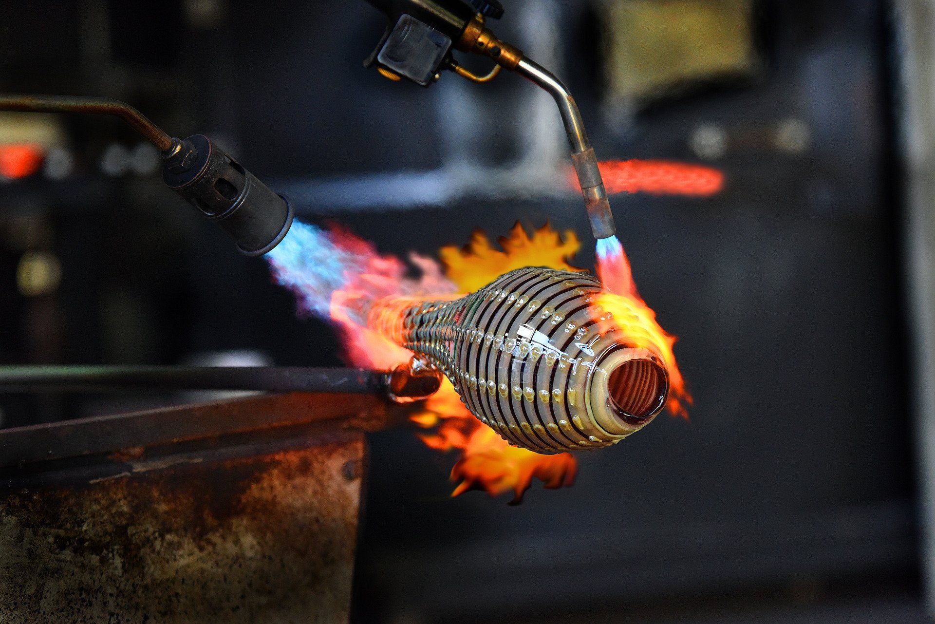 Glassblower shaping a ribbed glass vessel with a torch, surrounded by flames.
