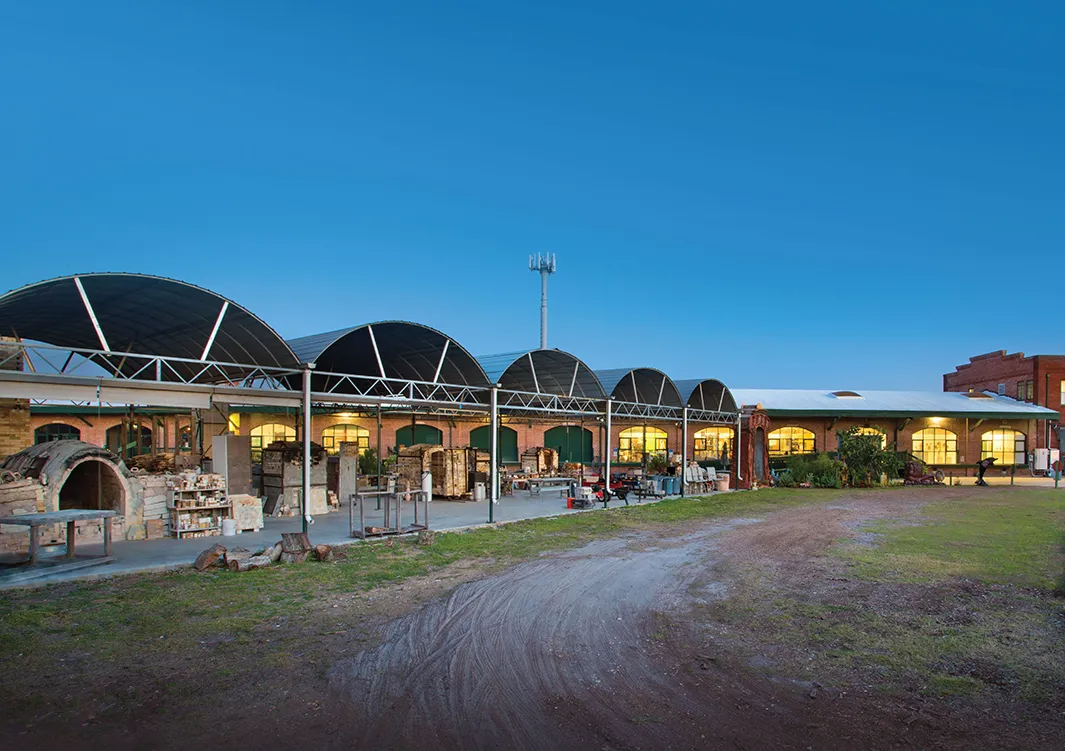 A long, low building with arched roofs, lit up at dusk, with a gravel path in front.