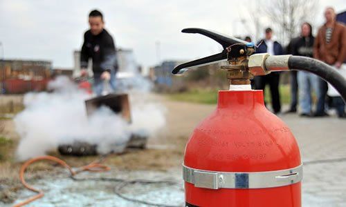 Extintor rojo en primer plano, persona apagando el fuego con una manguera. Un grupo observa.