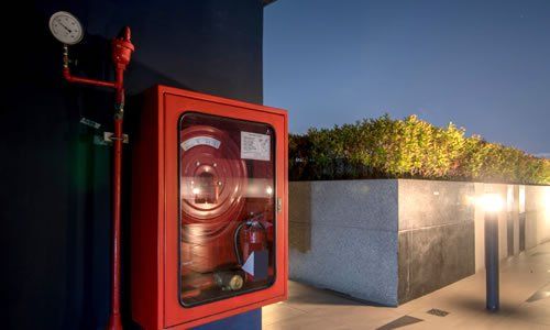 Armario rojo para manguera contra incendios sobre una pared azul oscuro con manómetro. En un jardín con iluminación exterior.