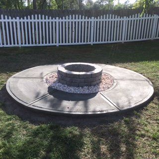 A fire pit is in the middle of a concrete patio in front of a white picket fence.