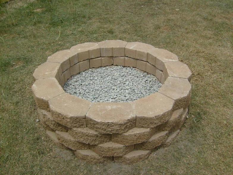 A fire pit made of bricks and gravel is sitting in the grass.