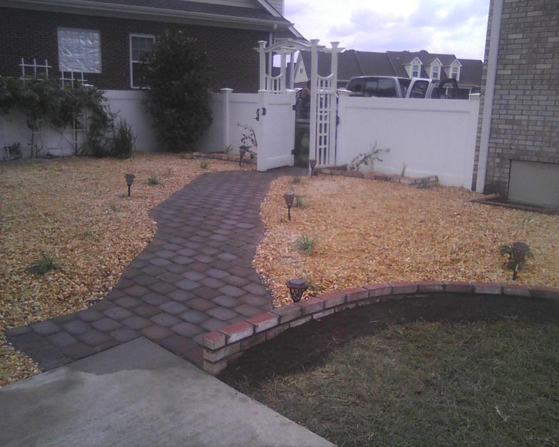 A brick walkway leading to a house with a white fence
