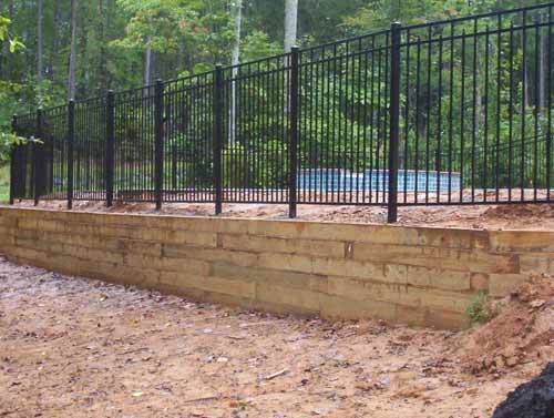 A wooden retaining wall next to a metal fence with a pool in the background.