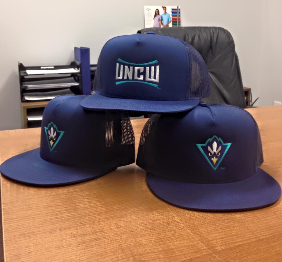 Three blue UNC Wilmington hats stacked on a wooden desk.