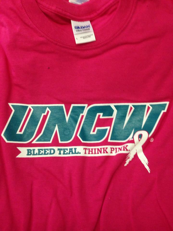 Pink UNCW t-shirt with teal and white lettering and a white ribbon for breast cancer awareness.
