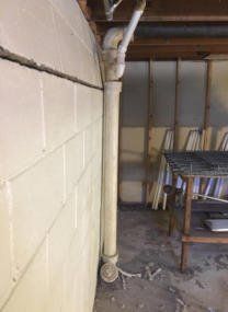 Basement corner with cinderblock wall, vertical pipe, and stacked boards beside a metal table Basement corner with cinderblock wall, vertical pipe, and stacked boards beside a metal table