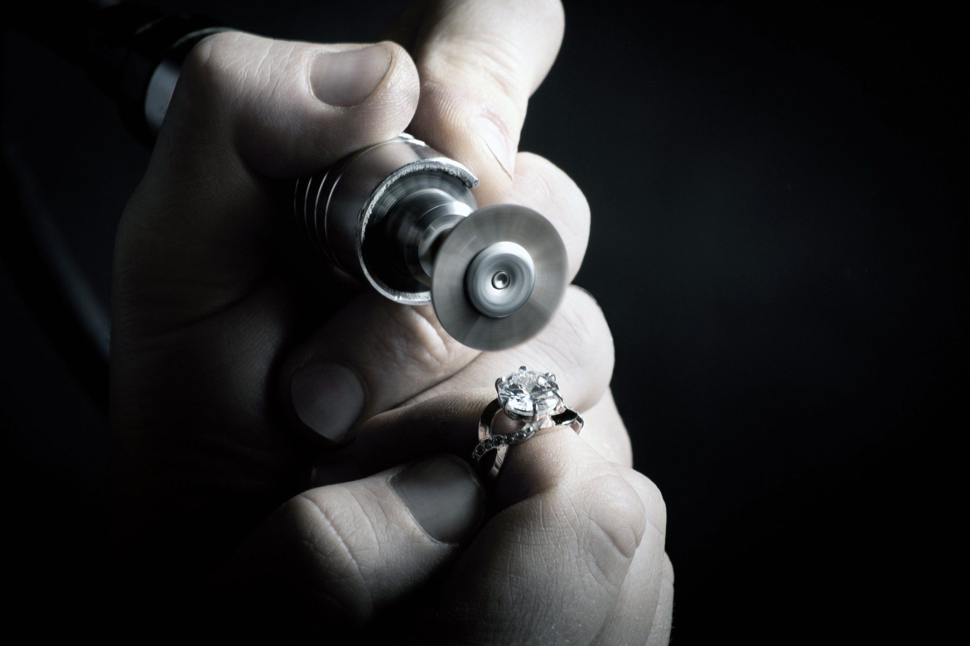 Jeweler polishes a diamond ring with a rotary tool, holding it against a black background.