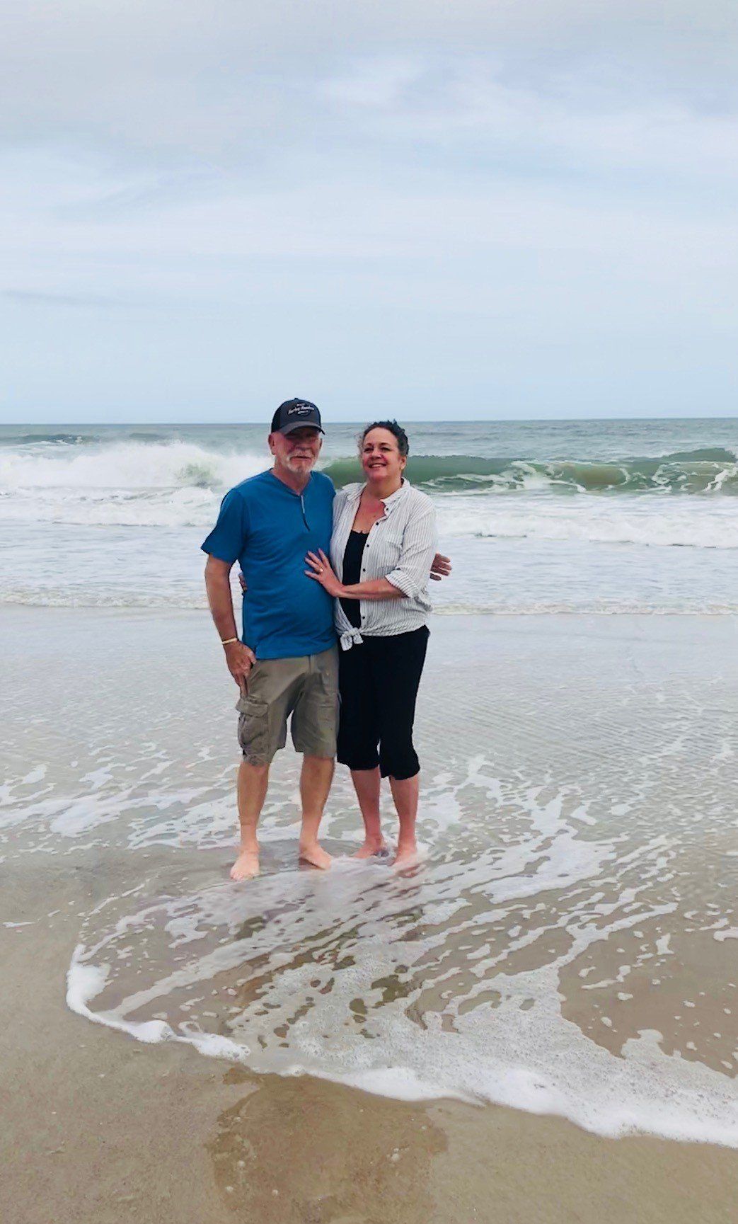 Man and woman standing at the beach, waves lapping at their feet. Cloudy sky, they are smiling and embracing.