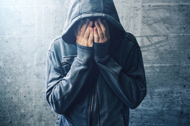 A man in a hoodie is covering his face with his hands.