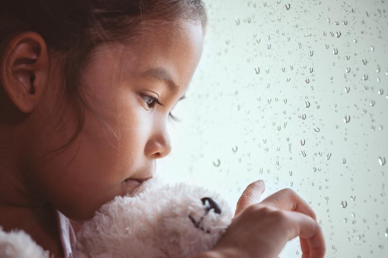 A little girl is holding a teddy bear and looking out of a window.