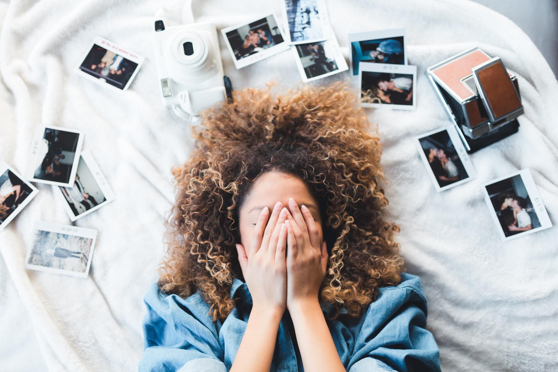 A woman is covering her face with her hands while laying on a bed surrounded by pictures.