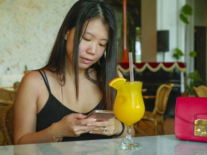 A woman is sitting at a table with a drink and looking at her phone.