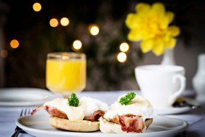 Eggs Benedict on a plate, with a glass of orange juice and a cup of coffee, with bokeh lights and a yellow flower in the background.