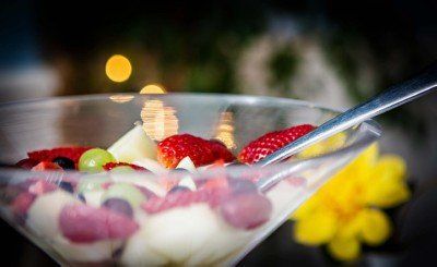 Fruit salad in a clear glass bowl with a spoon, containing strawberries, grapes, and other fruits, yellow flower in the background.