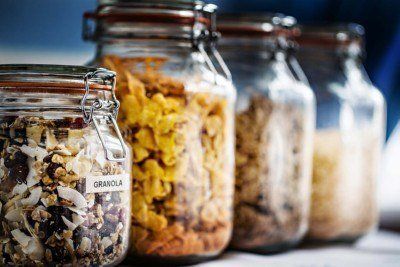 Four glass jars filled with various cereals, including granola, are lined up on a shelf, likely for storage.