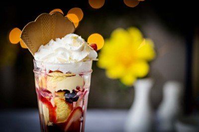Ice cream sundae in a glass, with whipped cream, a wafer cookie, and berries. Blurred background with lights and a yellow flower.