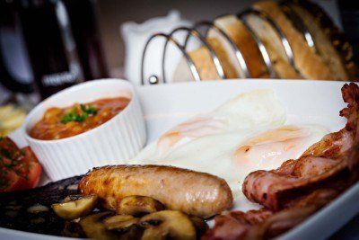 Full English breakfast on a white plate: eggs, bacon, sausage, beans, grilled tomato, mushrooms, and toast.