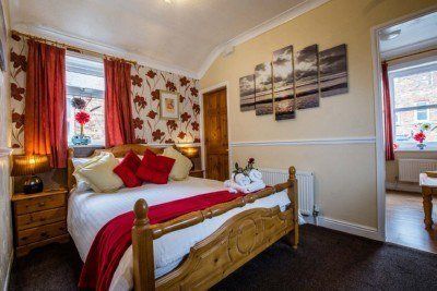 Cozy bedroom with a wooden bed, floral wallpaper, red curtains, and decorative pillows; a doorway leads to another room.
