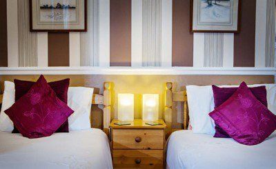 Twin beds with white linens, burgundy pillows, and a wooden nightstand with lit lamps, against a striped brown and white wall.