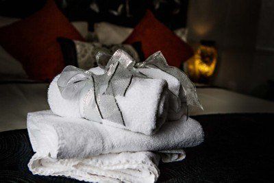 White towels, some rolled and tied with a silver ribbon, stacked on a bed with orange pillows.