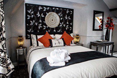 Elegant bedroom with a black and white floral headboard, bed with orange pillows, and a small table with a mirror and flowers.