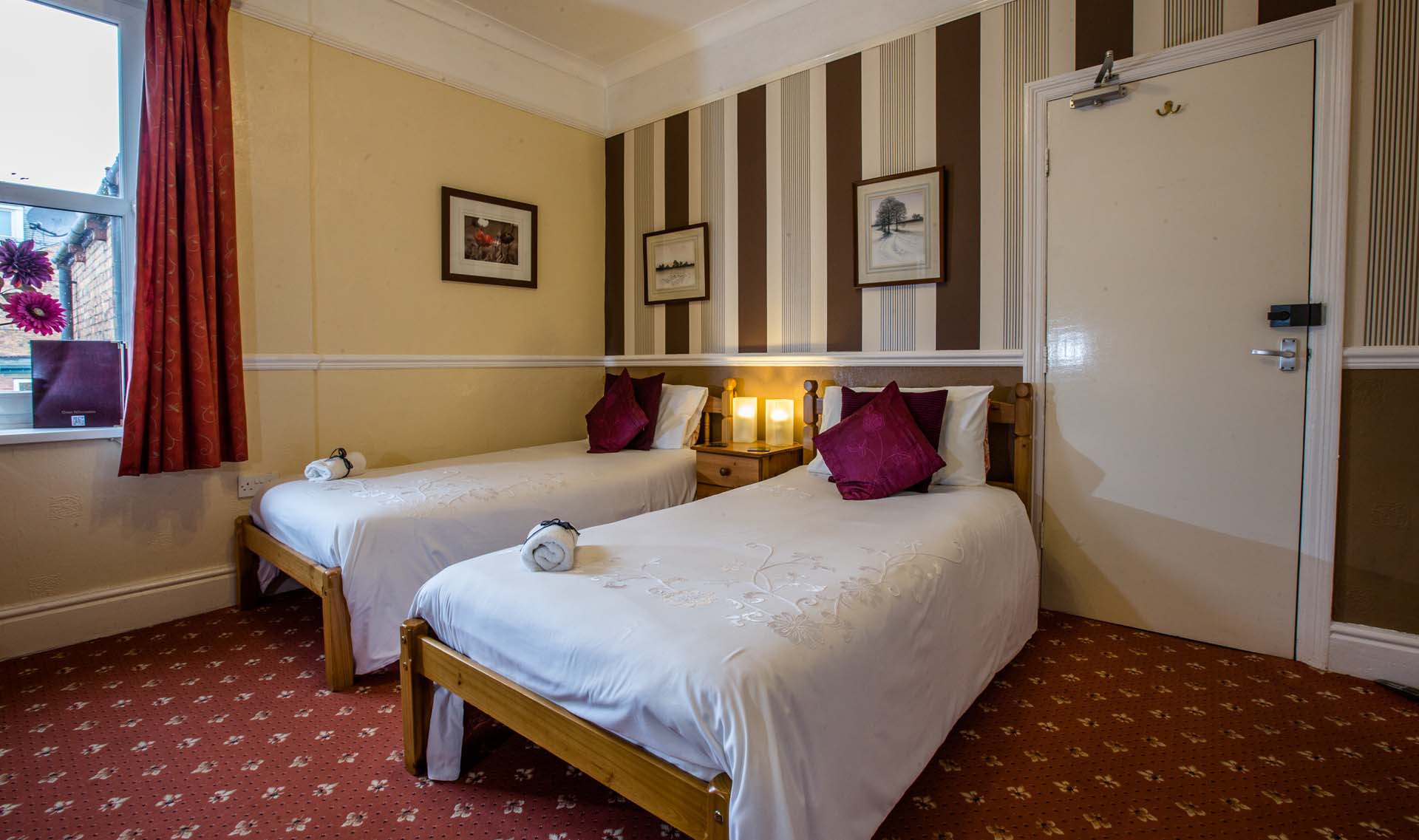 Twin hotel room with two beds, window with red curtains, and striped wallpaper.