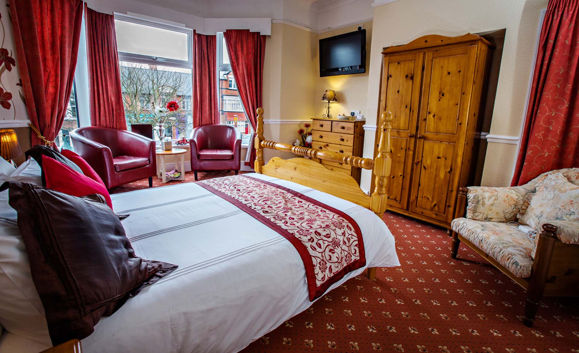 Cozy bedroom with a bed, armchairs, red curtains, and a large wooden wardrobe. Red patterned carpet and accents.