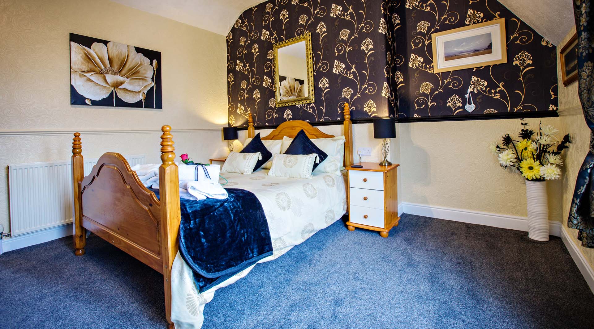 Bedroom with a wooden four-poster bed, dark patterned wallpaper, and a small white nightstand. The bed is made with white and blue bedding.