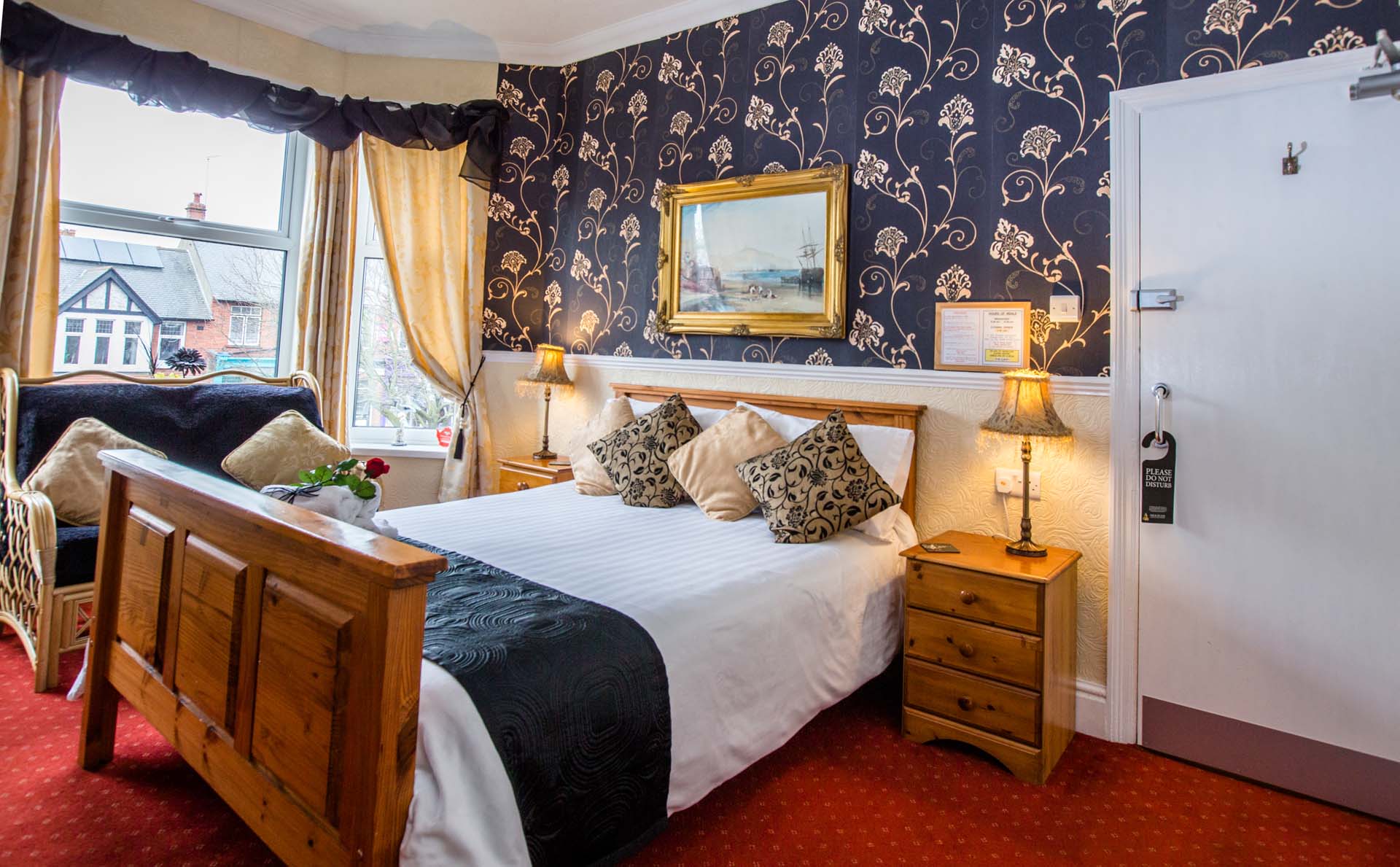 Cozy hotel bedroom with a bed, patterned wallpaper, and a bay window overlooking a street.