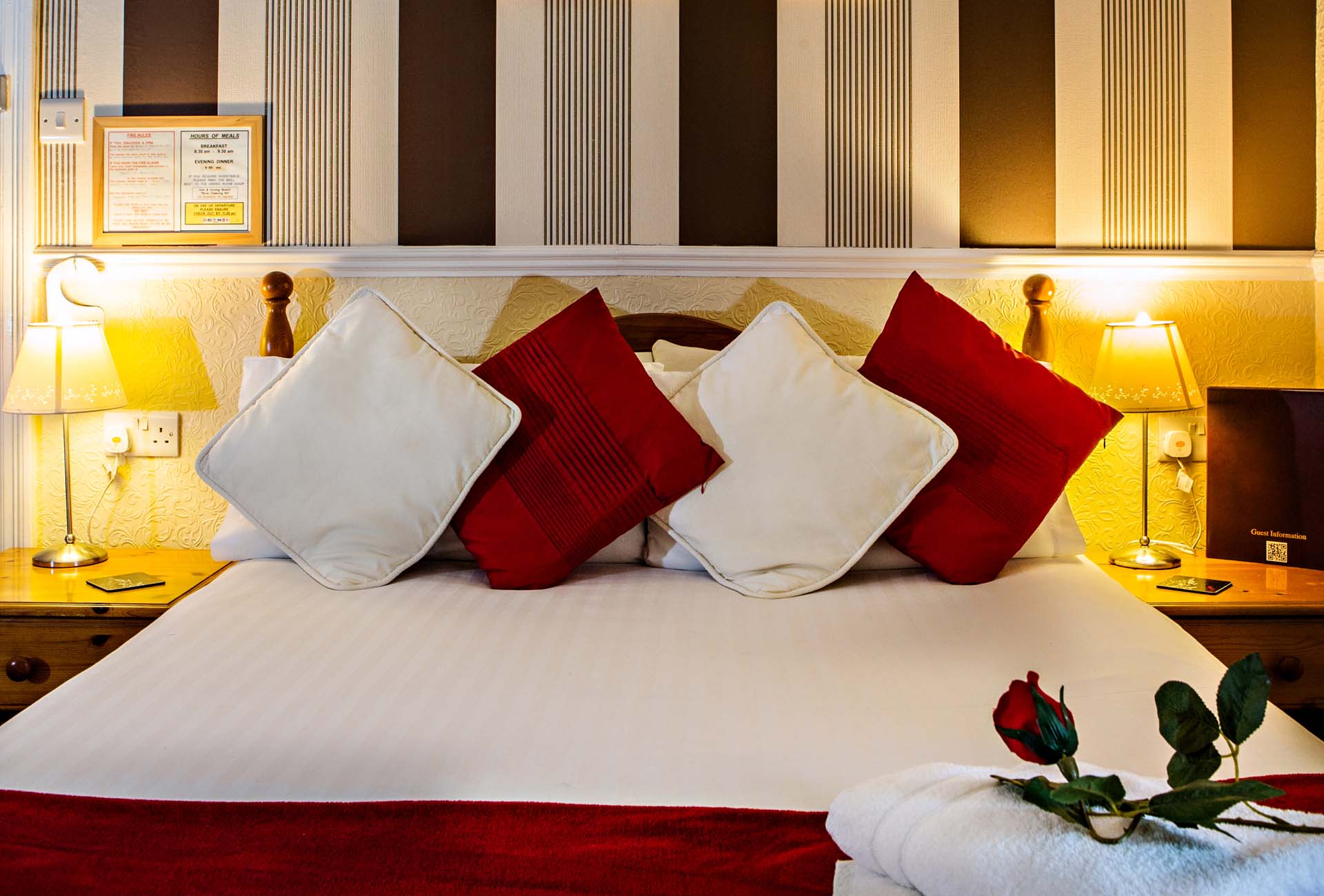 Bedroom with white and red pillows on the bed, nightstands with lamps, and a rose. Brown and white striped wallpaper is behind the bed.