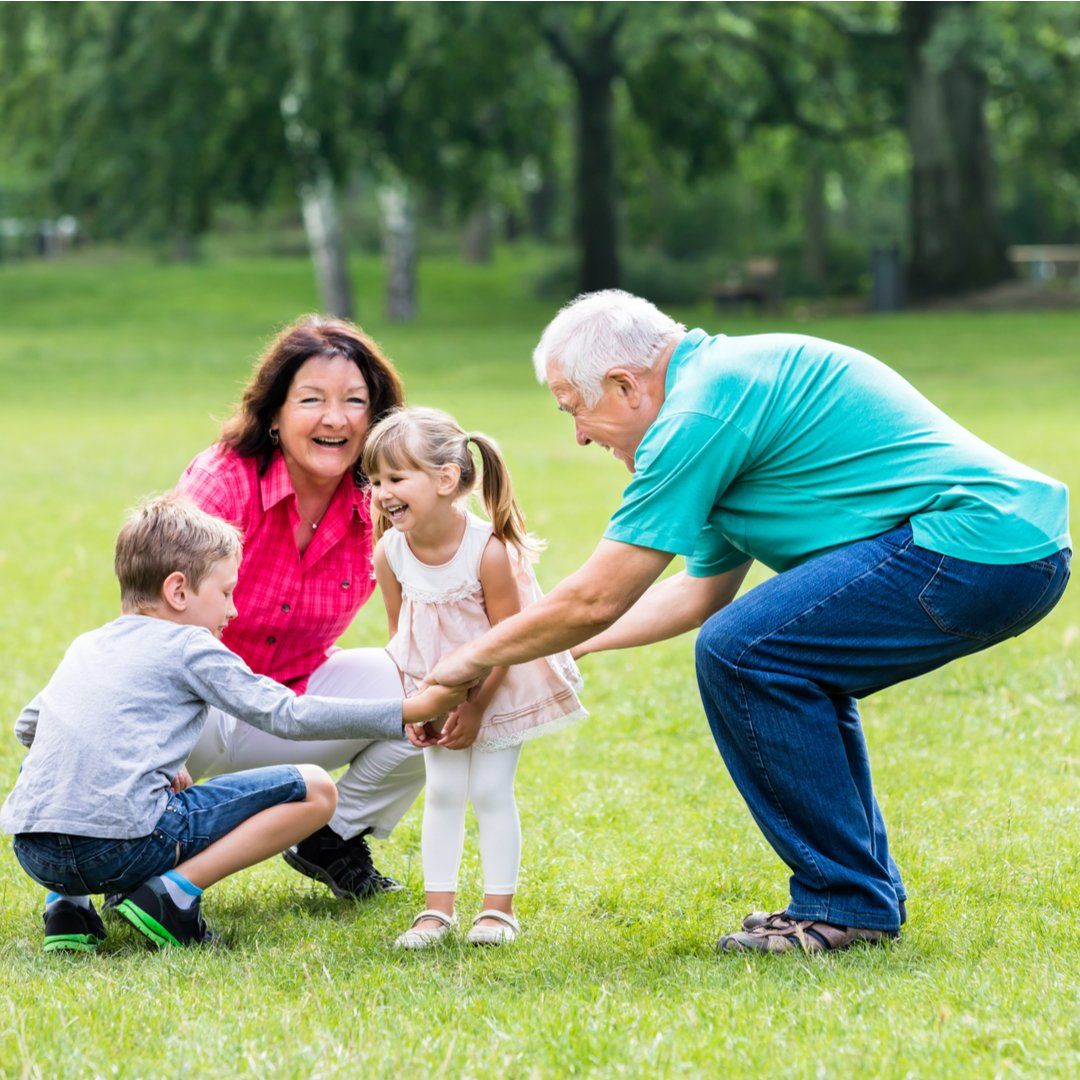 A group of four people interact playfully in a green park, with an older adult crouching to hold a young child’s hands.