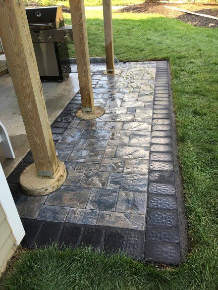 A stone walkway leading to a grill under a pergola in a backyard.