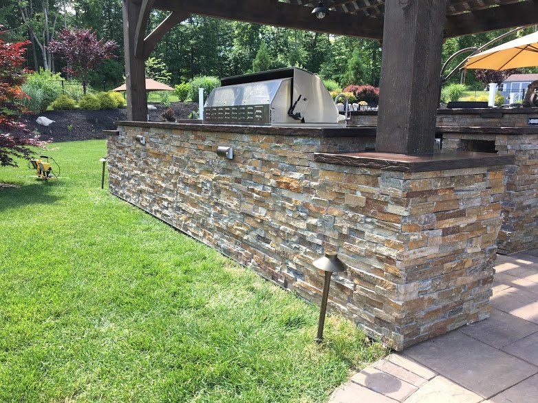 A stone wall with a grill under a pergola in a backyard.