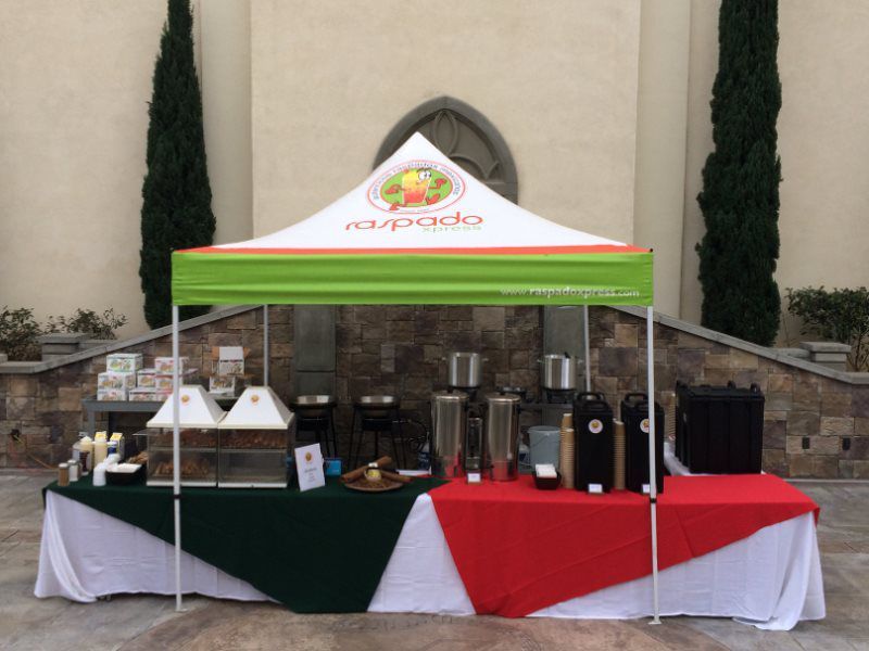 Food stand with green and red tables under a white tent with a logo, display of food items.