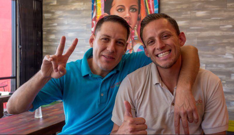 Two men smiling, one with a peace sign, the other with a thumbs up, arm around the other.