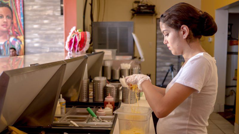 Woman in a white shirt, preparing food behind a counter in a restaurant.