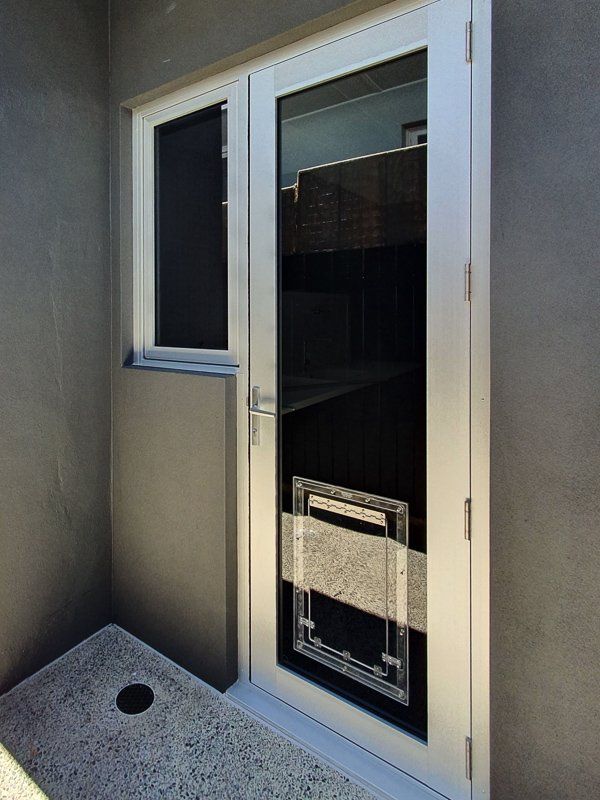 Glass door with built-in pet door and adjacent window, set in a gray wall, on a speckled concrete floor.