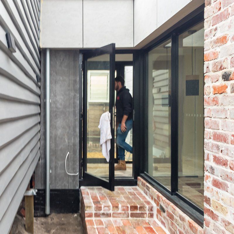 Man entering a building with glass doors. Red brick steps and a gray siding wall are visible.