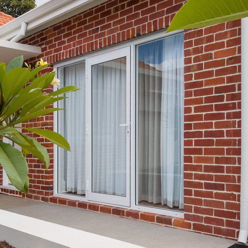 White-framed door and window with sheer curtains, set in a red brick wall. Green plant in the foreground.