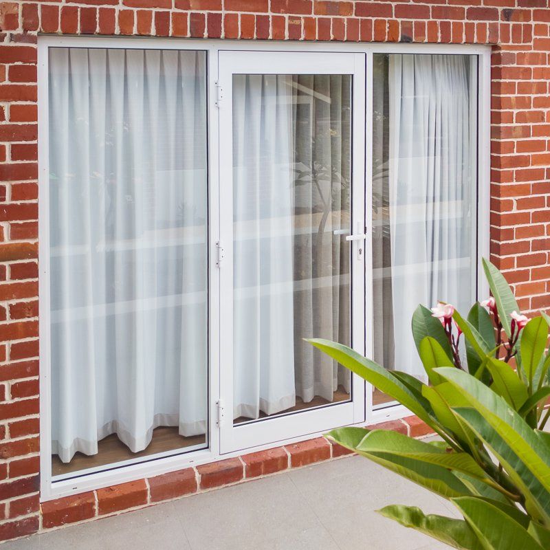 White-framed glass door and windows with white curtains set in a red brick wall, with a small plant in the foreground.