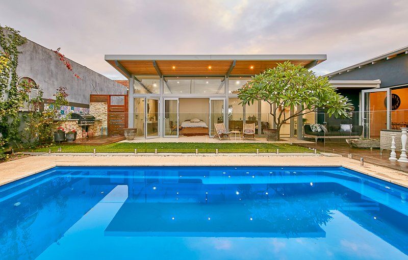 Backyard with pool, modern glass-walled extension, bedroom visible, lush green lawn, cloudy sky.