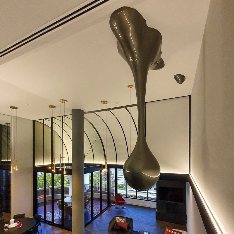 Sculptural art piece suspended from a high ceiling, above a modern living space with large windows and a balcony.